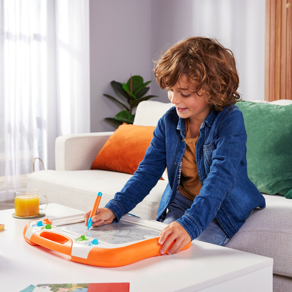 TOMY-style orange sketch board with pen stylus, used by a child on a couch
